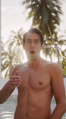 Vertical video portrait of shocked young man looking at camera and reacting on bad news, closes his mouth with his hand in horror, the emotion of fear, standing outdoors in resort hotel near pool