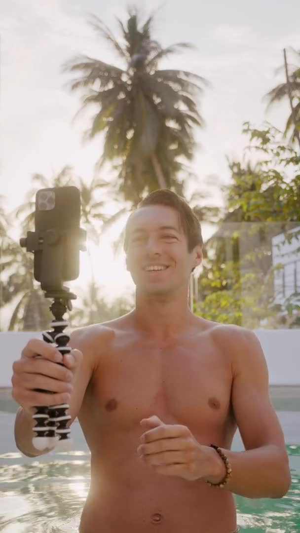 Vertical video of smiling young man holding mobile phone and making selfie on smartphone mounted on mini tripod while holidays outdoors at luxury pool villa at sunset — Wideo stockowe