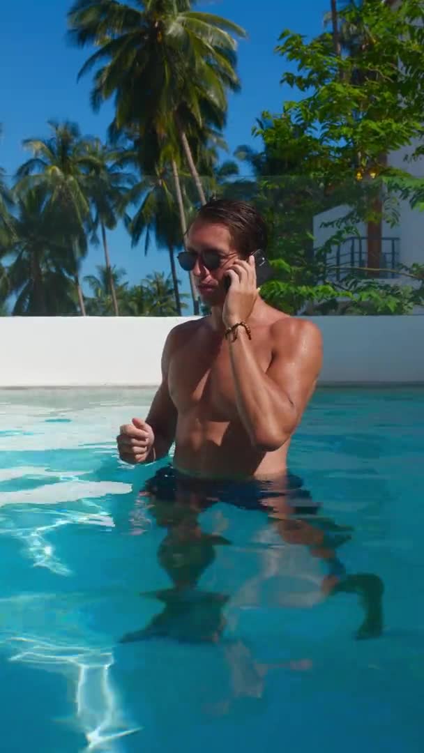 Vertical video portrait of a man in sunglasses talking on a mobile phone while standing in pool against the backdrop of palm trees. Male tourist talking on the phone on vacation — Wideo stockowe