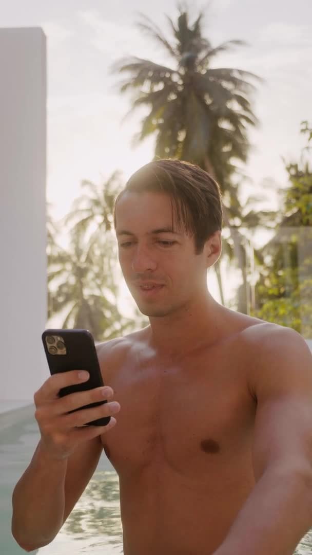 Vertical video portrait of smiling young man stands outdoors at hotel resort near pool and using app smartphone on beautiful sunset while holidays travel — Stock Video