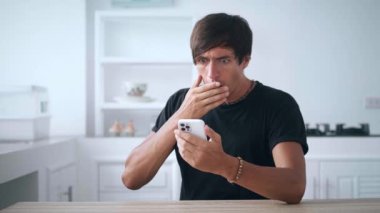 Şok olmuş genç adam akıllı telefon kullanıyor, kaybediyor, evdeki kötü haberlerden memnun değil. Üzgün erkek, arka plandaki mutfakta cep telefonunu tutuyor. Mobil kaybetme konsepti