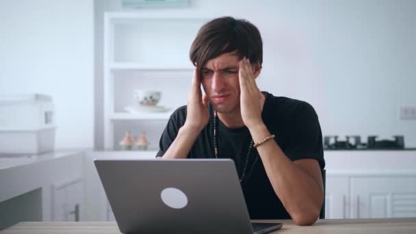 ラップトップで働いている間に頭痛に苦しんでいる悲しい男性のフリーランサーは、キッチンテーブルに座っている間の問題を心配しています。過労者の肖像自宅からノートパソコンでリモートで働いている. — ストック動画