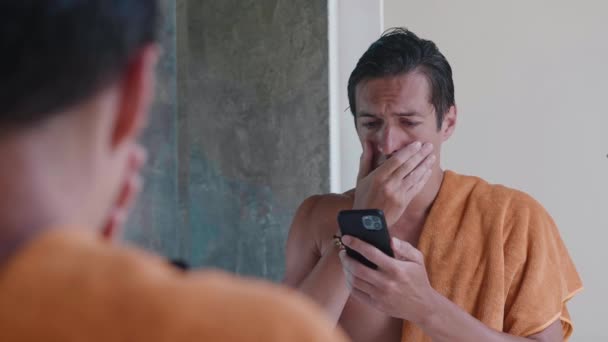 Σοκαρισμένος άνθρωπος κοιτάζοντας ένα smartphone και ανησυχούν για τα κακά νέα καλύπτει το πρόσωπό του με το χέρι του και κλαίει στη θλίψη και τη φρίκη, ενώ στέκεται στο μπάνιο — Αρχείο Βίντεο