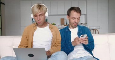Gülümseyen homoseksüel erkekler boş zamanlarını evdeki kanepede oturarak geçirirler. Bir adam dizüstü bilgisayar ve kulaklıkla müzik dinliyor, diğeri akıllı telefon kullanıyor. LGBT ilişkileri. 4K