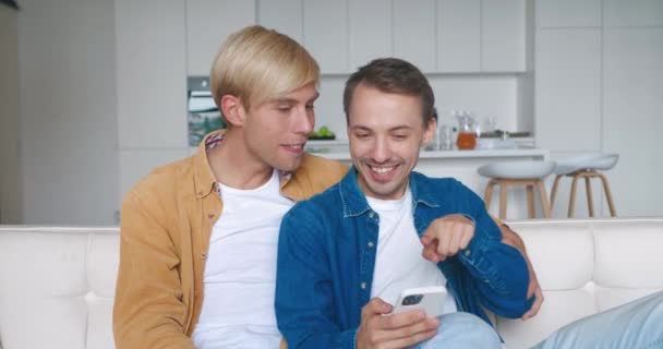 驚くべき2人の男性ゲイのカップルの勝者は ソファに座っている間に自宅でモバイルオンラインベット入札ゲームの勝利に驚かされた良いニュースをスマートフォンを保持します — ストック動画