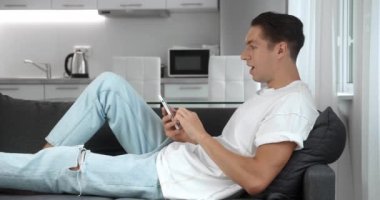 Mutlu genç adam iyi haberler alıyor. Heyecanlı erkek kazanan akıllı telefona bakarak başarıyı kutluyor ve evde kanepede dinlenirken evet jestleri yapıyor. Mobil zafer konsepti. 4K