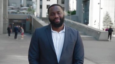 Takım elbiseli, kameraya bakan gülümseyen Afro-Amerikan profesyonel adam. Kendine güvenen yakışıklı iş adamı, lider, şehir merkezinde, iş merkezinin yakınında poz veren bir yönetici. 4K