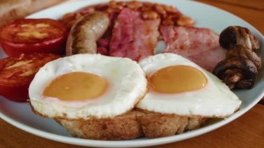 English breakfast close up - fried eggs, sausages, bacon, tomatoes, tomato beans, toasts - in white plate on wooden table. High quality 4k footage