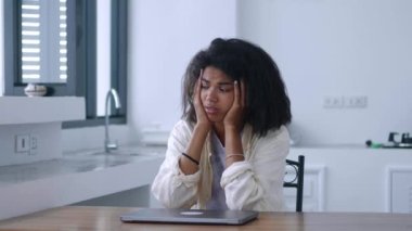 Young worried woman sitting at a table with a laptop, looking aside with despair and feeling depressed and unmotivated.
