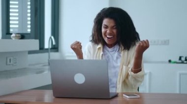 Successful harworking millennial young woman copywriter, clenching fists and rejoicing while working on laptop from home