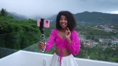 Charming young African woman smiles while recording video or talking on the phone by video call standing on her balcony