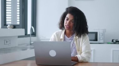 Confident young hardworking multitaskin African woman, freelance worker, sitting on a wooden desk, during a video call