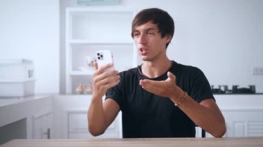 Akıllı telefonu tutan ve cep telefonuyla konuşan genç bir adam, evde, mutfakta. 4K