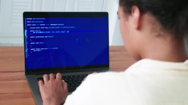 Rear view of a programmer, using laptop, writes computer programs, sitting on a wooden desk at home office interior.