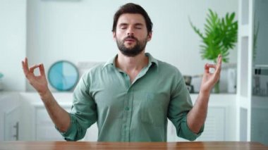 Koyu renk saçlı, çoklu etnik kökenli bir adam ev mutfağında bir masada tek başına oturuyor ve lotus pozisyonunda meditasyon yapıyor.