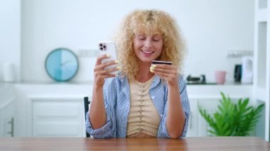 Güzel kıvırcık saçlı sarışın kadın internet alışverişini seviyor, akıllı telefonunu kullanıyor, evden internet üzerinden ödeme yapıyor.
