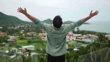 Terasta duran, elini kaldıran, gökyüzüne minnettar bir şekilde bakan, deniz kenarının güzel manzarasına hayran bir adamın arka görüntüsü.