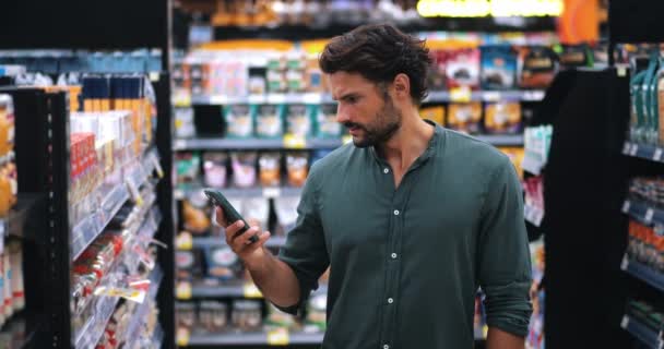 魅力的な白人男性は彼のスマートフォンを使用しています 食品スーパーマーケットで買い物中に購入する製品のリストをチェック — ストック動画