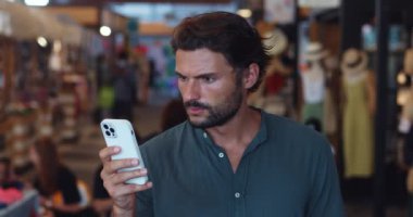 Beyaz adam akıllı telefon kullanıyor, haber akışını kaydırıyor, şok geçiriyor, tanınmayan bir alışveriş merkezinde duruyor.