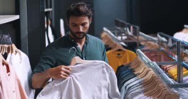 Çekici sakallı adam alışverişçisi, erkek giyim kuşamlarını denetliyor, moda bir alışveriş merkezinde elbise askılarında asılı duruyor..
