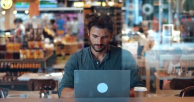 Yoğunlaşmış genç adam, paket servis kupasından kahve içiyor, dizüstü bilgisayar kullanıyor, yeni bir proje ve iş stratejisi planlıyor.