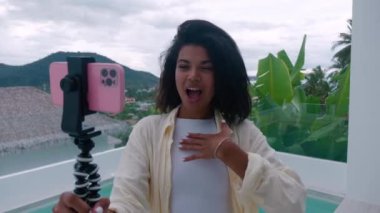 Güzel bir kadın içeriği kaydediyor. Kanal için vlog, balkonda havuz başında duruyor ve arka planda inanılmaz tropik bir köy var.