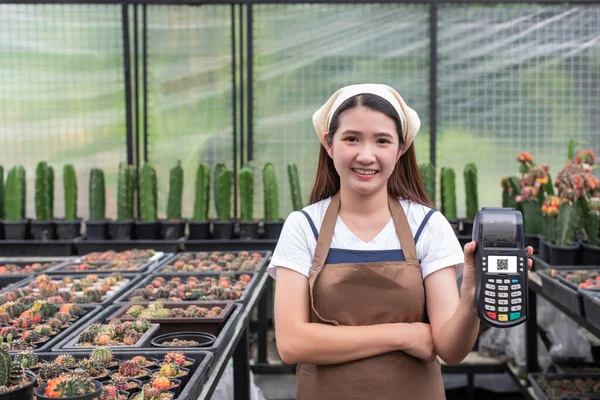 Smiling young asian woman agriculture employee in apron hold modern bank payment terminal to process and acquire credit card payments  on green house cactus farm background.