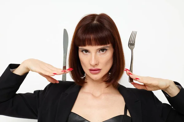 Beautiful woman in a black jacket a knife and a fork near the face light background — Stockfoto