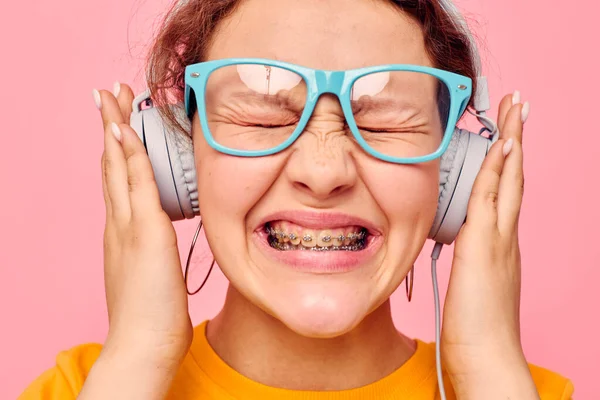 Mavi gözlüklü komik kız kulaklıkla müzik dinliyor. Pembe arka plan değiştirilmemiş. — Stok fotoğraf
