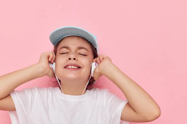 Portrait of happy smiling child girl headphones in a white t-shirt and a cap pink background — Stock Fotó