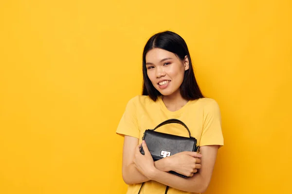 Rolig asiatisk kvinna i en gul t-shirt svart kvinnors väska i händerna poserar — Stockfoto
