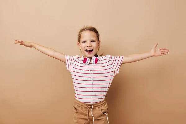 Маленькая девочка в полосатых наушниках на футболках жестом руки бежевый фон — стоковое фото