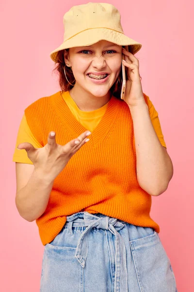 Funny girl in an orange sweater in yellow hat talking on the phone pink background unaltered — Stockfoto