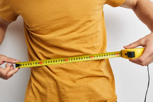 Handsome guy measuring tape measure in yellow t-shirt light background — Stock Photo, Image