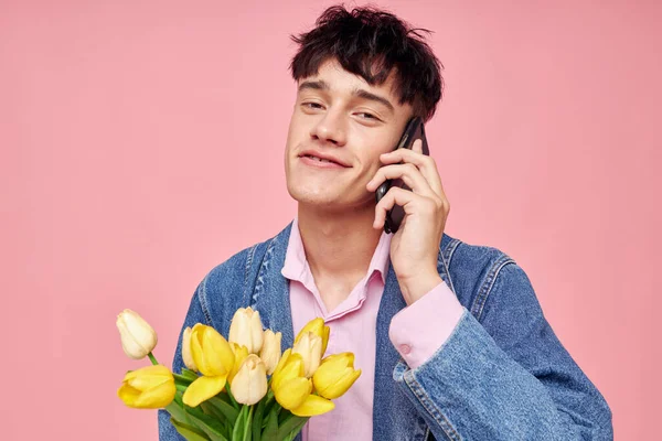 若い男がデニムジャケットに花の花束を携帯電話のピンクの背景で話している — ストック写真