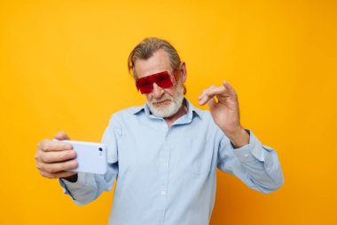 Yaşlı adam, elinde telefon, sarı arka planda duygularla poz veriyor.
