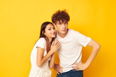 Beyaz tişörtlerle yan yana duran genç kız ve erkek sarı arkaplan pozu veriyor.