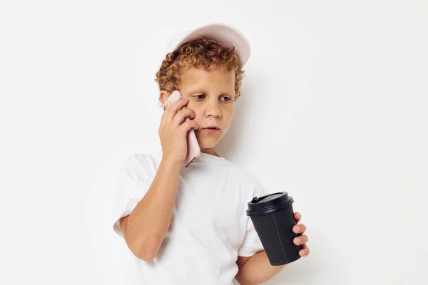 Niño pequeño con una gorra de camiseta blanca con un teléfono en un vaso con un fondo claro bebida inalterado — Foto de Stock