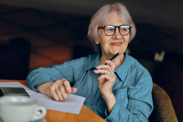 Anciana con gafas se sienta en una mesa frente a un portátil inalterado — Foto de Stock