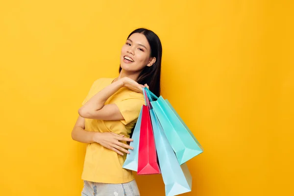 Vrouw met aziatische verschijning in een geel T-shirt met veelkleurige boodschappentassen geïsoleerde achtergrond ongewijzigd — Stockfoto