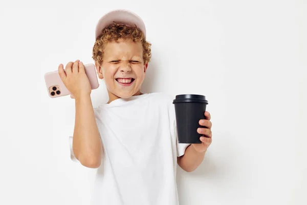 Foto retrato niño rizado en una gorra de camiseta blanca con un teléfono en un vaso con un fondo claro bebida inalterado — Foto de Stock