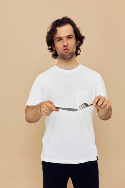 Hombre guapo en una camiseta blanca con cuchillo con tenedor de fondo beige —  Fotos de Stock