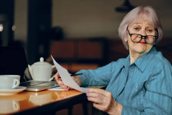 Mulher idosa com óculos senta-se em uma mesa na frente de um laptop Freelancer funciona inalterado — Fotografia de Stock