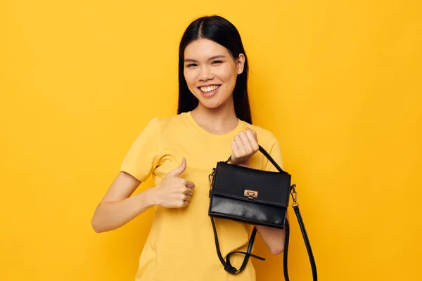 Charmig ung asiatisk kvinna med en svart kvinna väska i händerna gul bakgrund oförändrad — Stockfoto