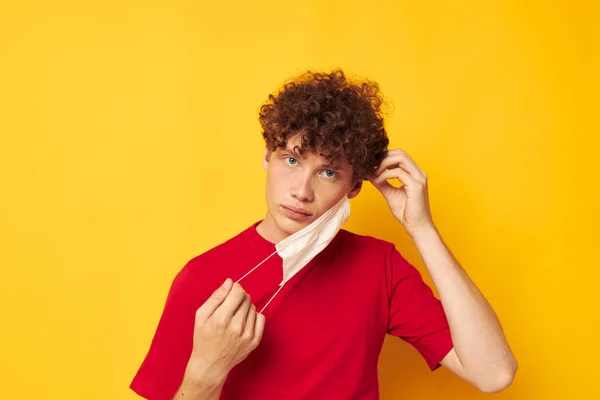 Tipo con el pelo rizado rojo en una camiseta roja máscara médica protección fondo amarillo inalterado — Foto de Stock