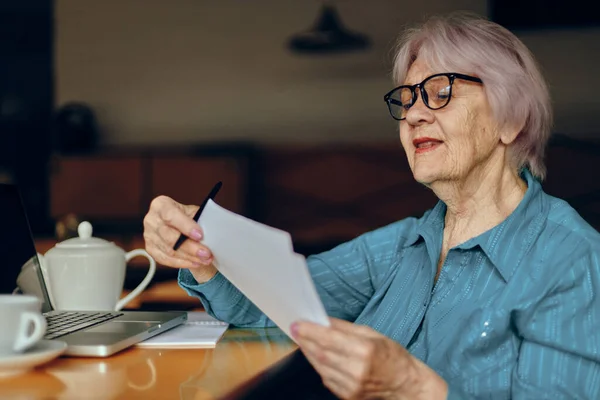Mujer mayor con gafas se sienta en una mesa delante de un ordenador portátil Freelancer trabaja sin alteraciones — Foto de Stock
