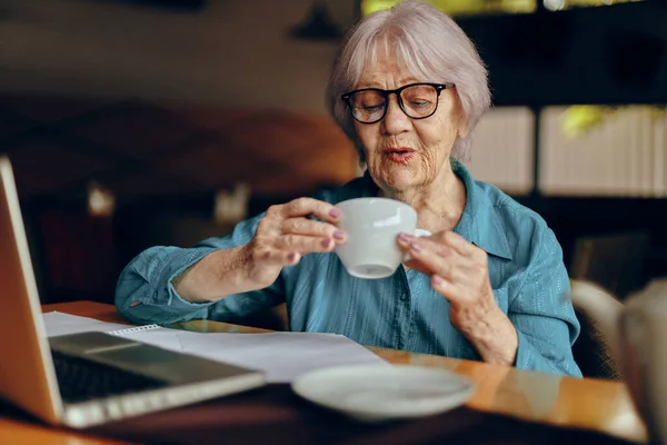 Happy senior woman with glasses sits at a table in front of a laptop Lifestyle unaltered — Stock Photo, Image