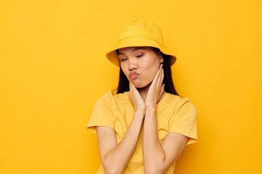 Sarı tişörtlü ve şapkalı çekici genç Asyalı kadın duygularının sarı arka planını değiştirmemiş.