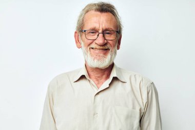 Gri sakallı, gömlekli, gözlüklü, açık renk arka planlı yaşlı, kır saçlı bir adam.