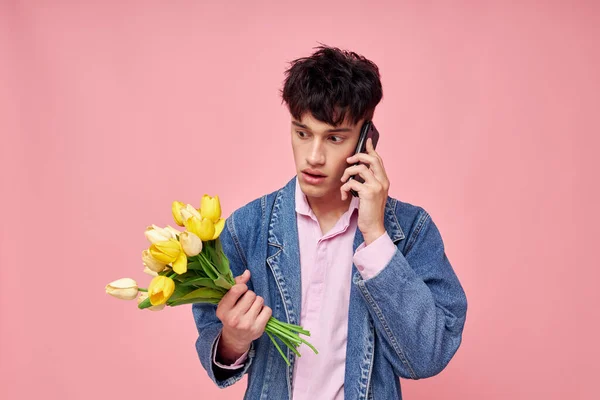 Молодой парень в джинсовой куртке с букетом цветов разговаривает по телефону розовый фон — стоковое фото